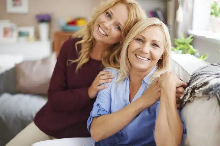 Two women sit closely together on a sofa, smiling at the camera in a bright, cosy living room—capturing the perfect moment for a memorable Mother’s Day present.