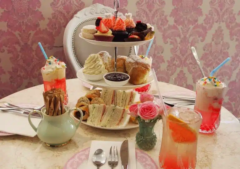 A tiered tray with sandwiches, scones, and desserts sits on a table set for tea with milkshakes, a teapot, cutlery, serviettes, and a small vase of pink roses—perfect as a thoughtful Mother's Day present.