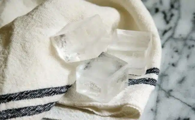 Three clear ice cubes rest on a white cloth with black stripes, placed on a grey marble surface—a simple, soothing scene that evokes the clarity many seek when tackling adult acne.