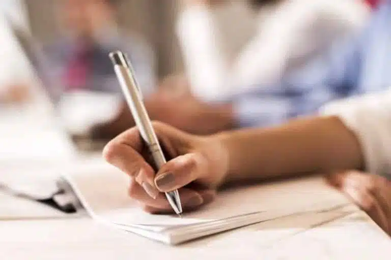 A hand holding a pen writes on a notepad at a desk during a gss 2017 meeting, with blurred people and a laptop in the background.