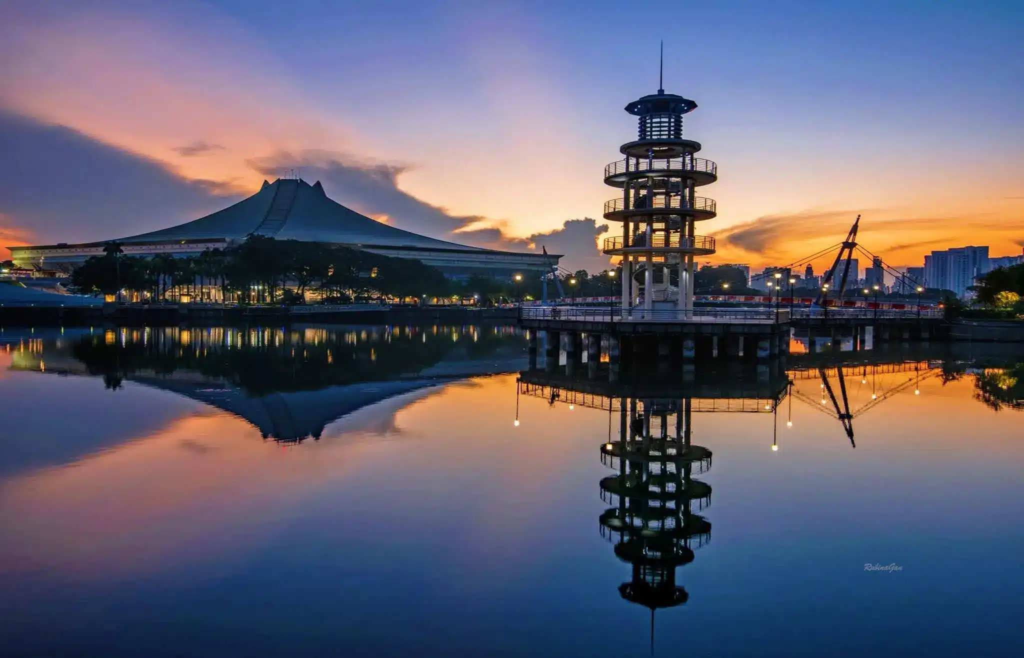 A modern tower and a domed structure are reflected in a calm lake at sunset, with orange and blue hues in the sky, capturing the tranquil atmosphere just before the NDP Fireworks light up the evening.
