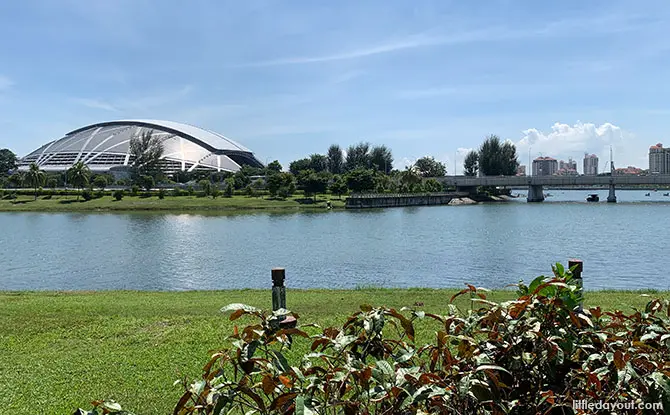 Singapore Sports Hub dome on the left, a bridge over the Kallang Basin, with blue sky, greenery, and city buildings visible in the background—an iconic view often illuminated by dazzling NDP fireworks during National Day celebrations.