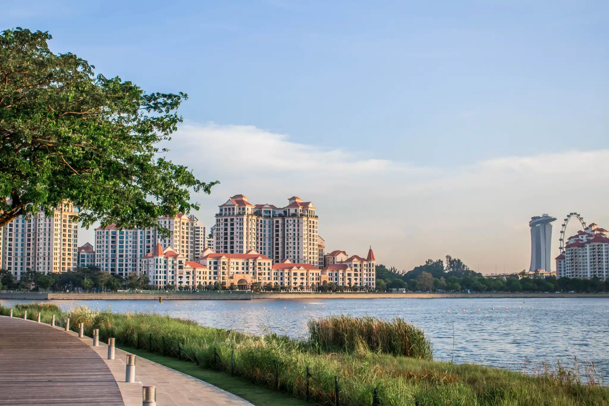 A riverfront walkway with grass, high-rise buildings in the background, a Ferris wheel, and Marina Bay Sands under a blue sky, offering a perfect vantage point to watch the spectacular NDP Fireworks illuminate the skyline.