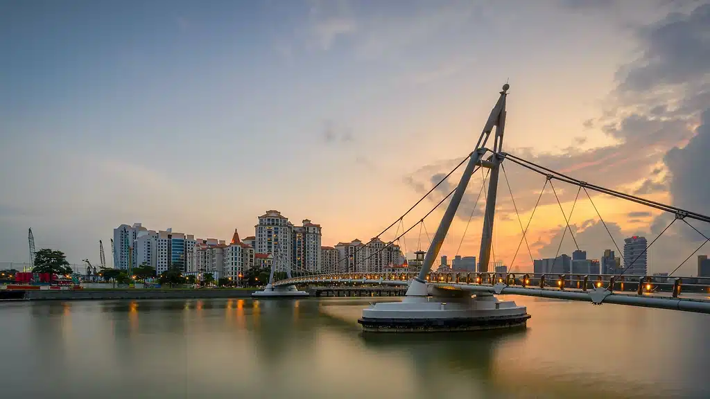 A modern pedestrian suspension bridge stretches over calm water with city buildings in the background at sunset, whilst bursts of NDP Fireworks illuminate the evening sky.