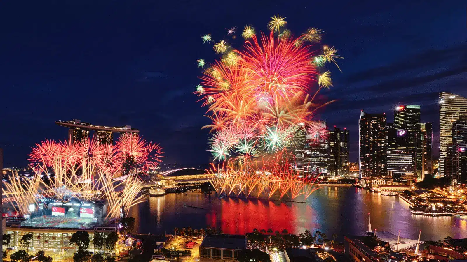 Colourful NDP Fireworks explode over a city skyline at night, reflecting on the water and illuminating surrounding buildings and landmarks.