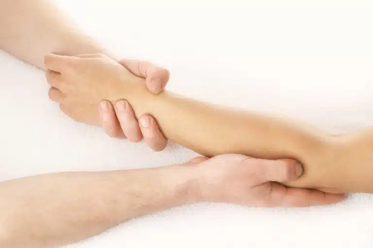 A person holds and gently massages another person's forearm and wrist, demonstrating a soothing Massage Therapy technique while both arms rest on a white surface.