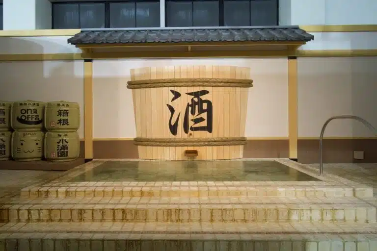 A large wooden barrel marked with Japanese kanji stands in a tiled indoor Sake Bath area, with water and steps leading into the pool. Sake barrels are stacked in the background, enhancing the authentic atmosphere.