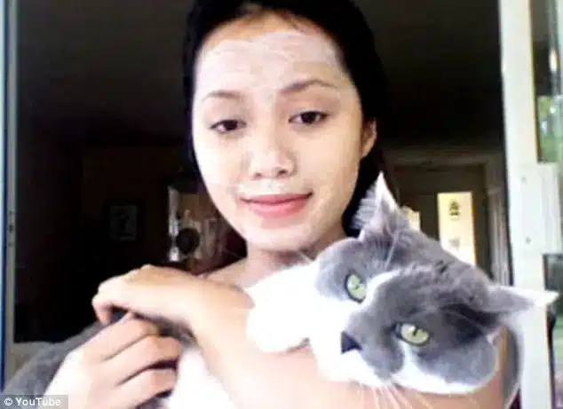 A woman with a white Japanese facial mask holds a grey and white cat in her arms, standing indoors near a doorway.