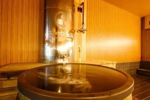 A large wooden tub filled with hot water sits in a room with wood-panelled walls and a metal water heater in the background, evoking the relaxing atmosphere of a traditional beer onsen.