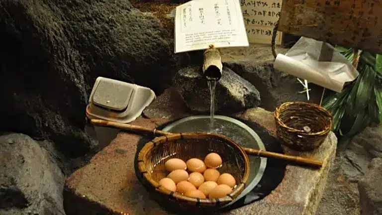 A bamboo spout pours water into a stone basin holding a basket of eggs, evoking the tranquil atmosphere of a beer onsen, with baskets and papers nearby in a natural setting.