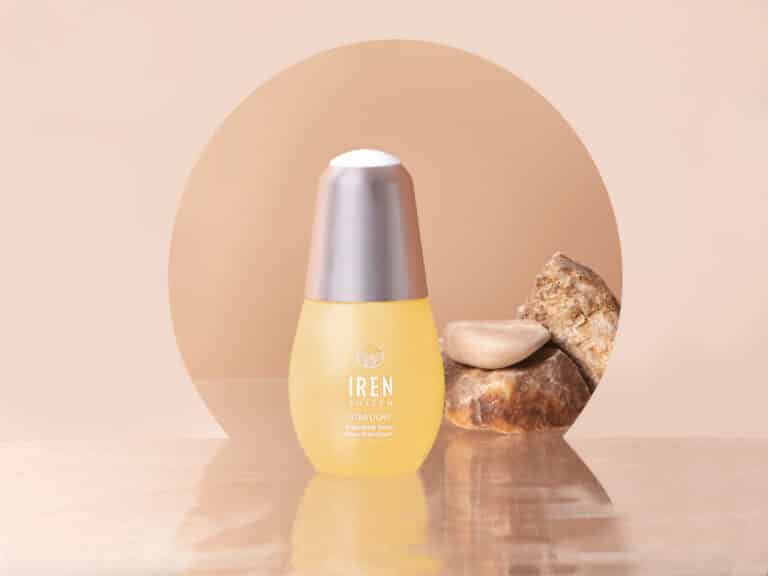 Yellow skincare serum bottle with a silver cap labeled "IREN Shizen" sits in front of rocks on a reflective surface, reminiscent of the allure of free beer, against a beige background.
