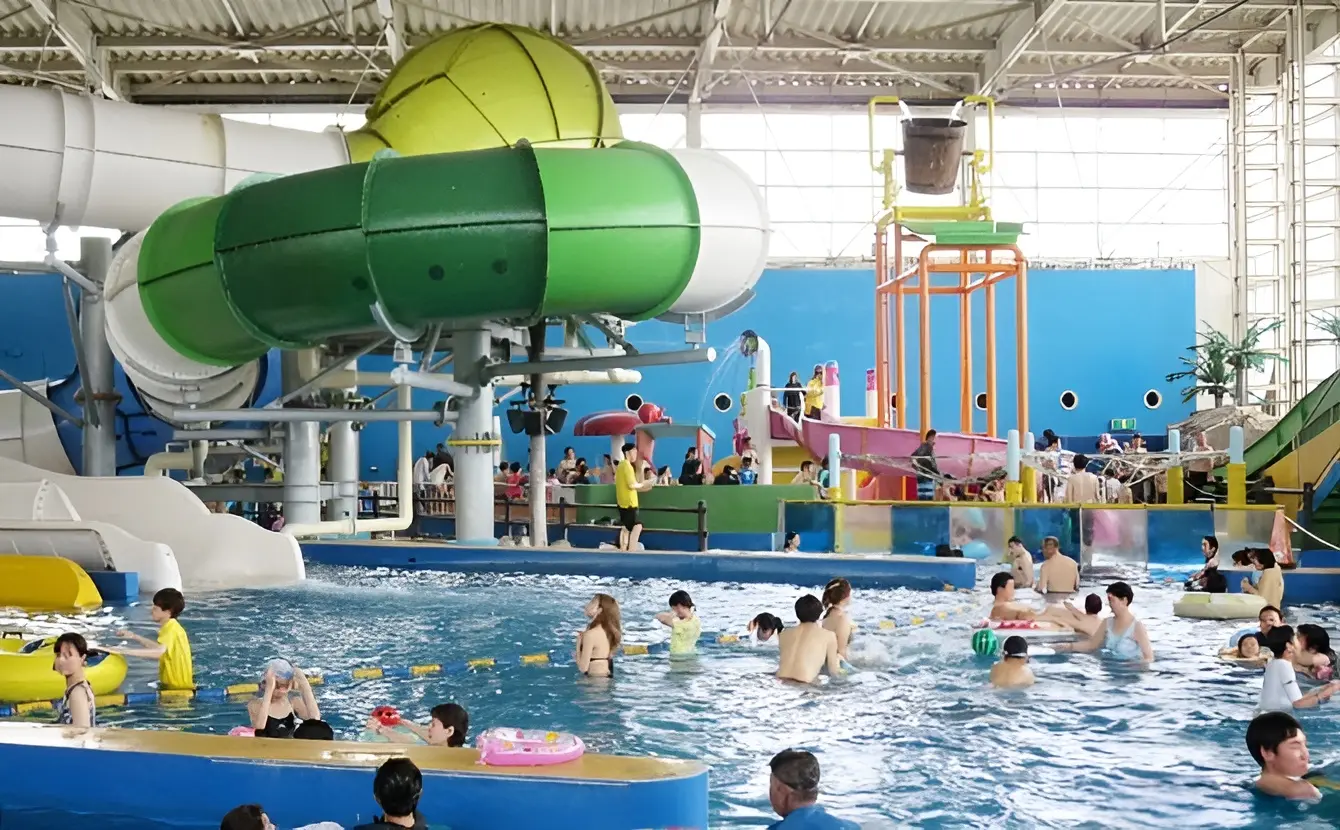 Indoor water park with people swimming in a pool, a large green waterslide, play structures in the background, and a relaxing mixed gender onsen area for guests to unwind after enjoying the attractions.