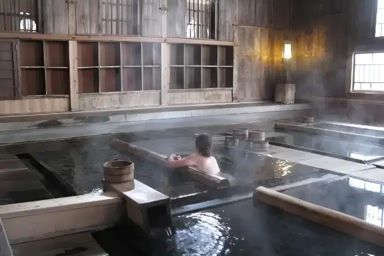 A person sits alone in a steaming indoor wooden mixed-gender onsen bath, with empty wooden shelves and buckets nearby.