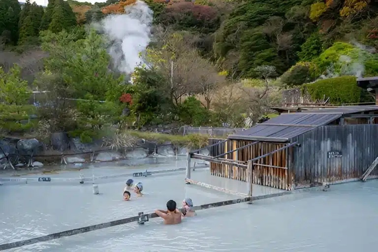 People bathe in a mixed gender onsen, soaking in outdoor hot springs with milky blue water, surrounded by greenery and steam rising in the background.