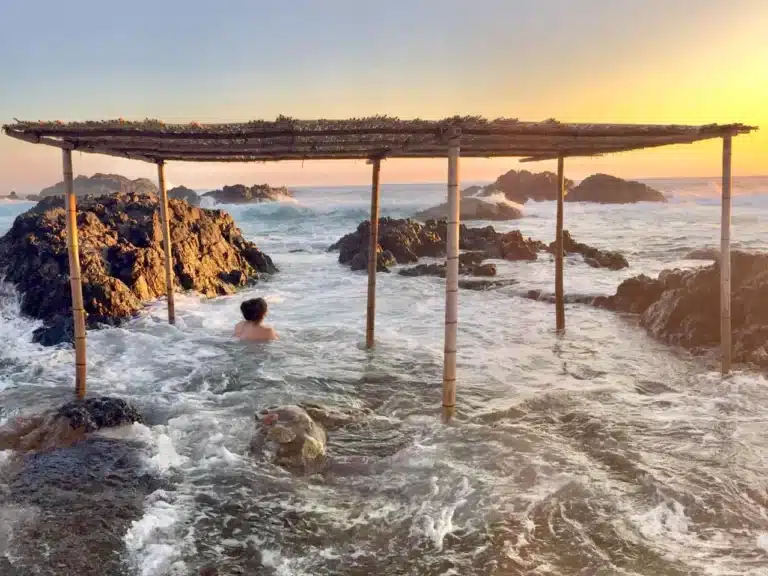 A person sits in shallow sea water beneath a simple bamboo shelter, reminiscent of a mixed-gender onsen, surrounded by rocks at sunset.
