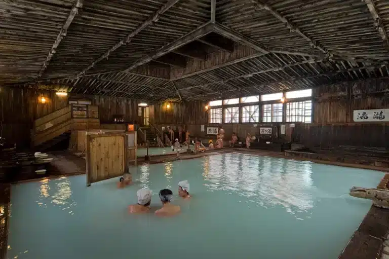People soak in a large indoor mixed-gender onsen with milky water, surrounded by rustic wooden beams and Japanese signage on the walls, creating an authentic and inviting atmosphere.