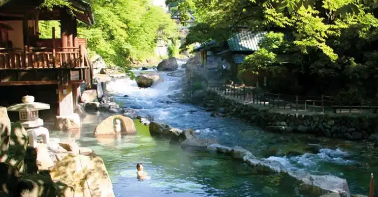 A person bathes in a mixed-sex onsen outdoor hot spring pool beside a flowing river, surrounded by lush greenery and traditional Japanese buildings.