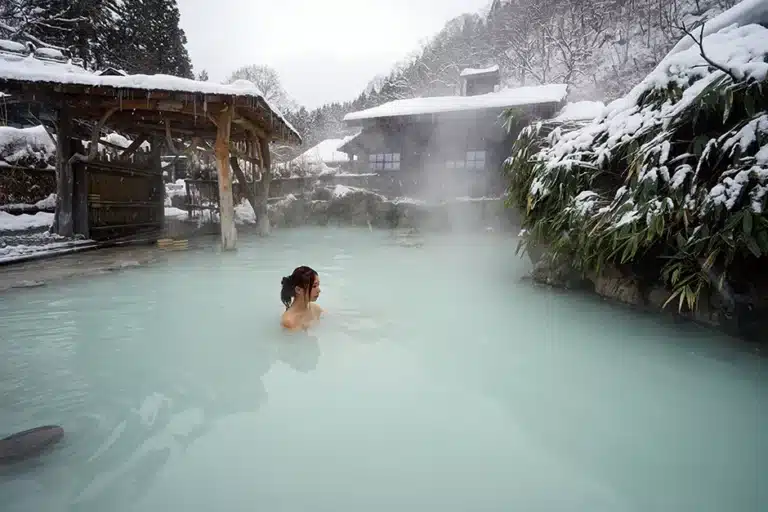 A person bathes in a mixed-gender onsen, an outdoor hot spring surrounded by snow-covered structures, trees, and mountains on a winter’s day. Steam rises from the milky blue water.