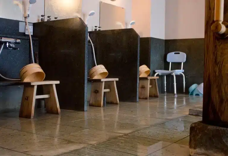 Wooden stools and buckets are arranged at individual washing stations in a tiled Japanese onsen bathhouse, with hand showers and mirrors on the walls.