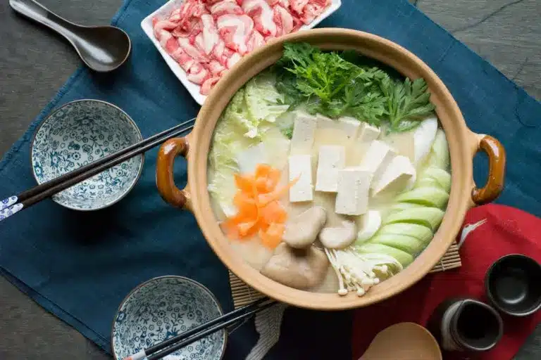 A pot of Japanese hotpot with tofu, mushrooms, greens, carrots, and sliced vegetables—perfect for learning how to keep warm in winter—surrounded by two bowls, chopsticks, and a plate of sliced meat.