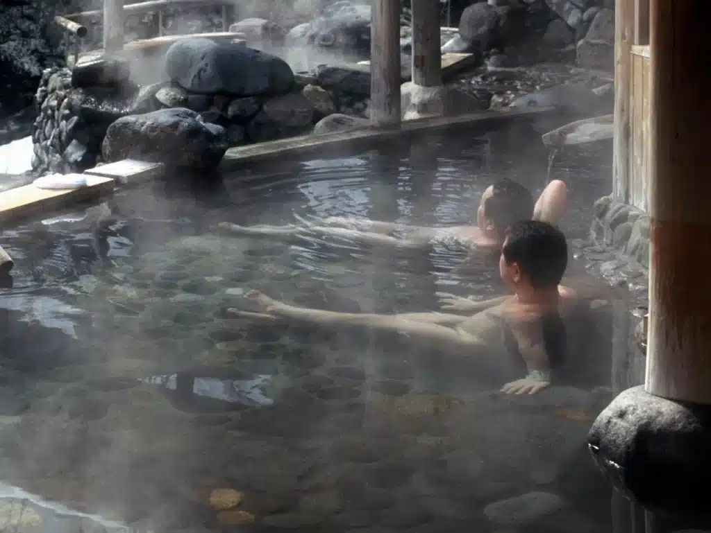 Two people relax in a steamy outdoor Japanese onsen, surrounded by rocks and wooden posts.