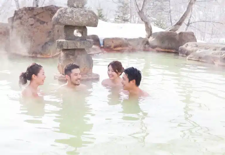 Four people relax and converse in an outdoor Japanese onsen surrounded by rocks and light steam, with snow and bare trees in the background.