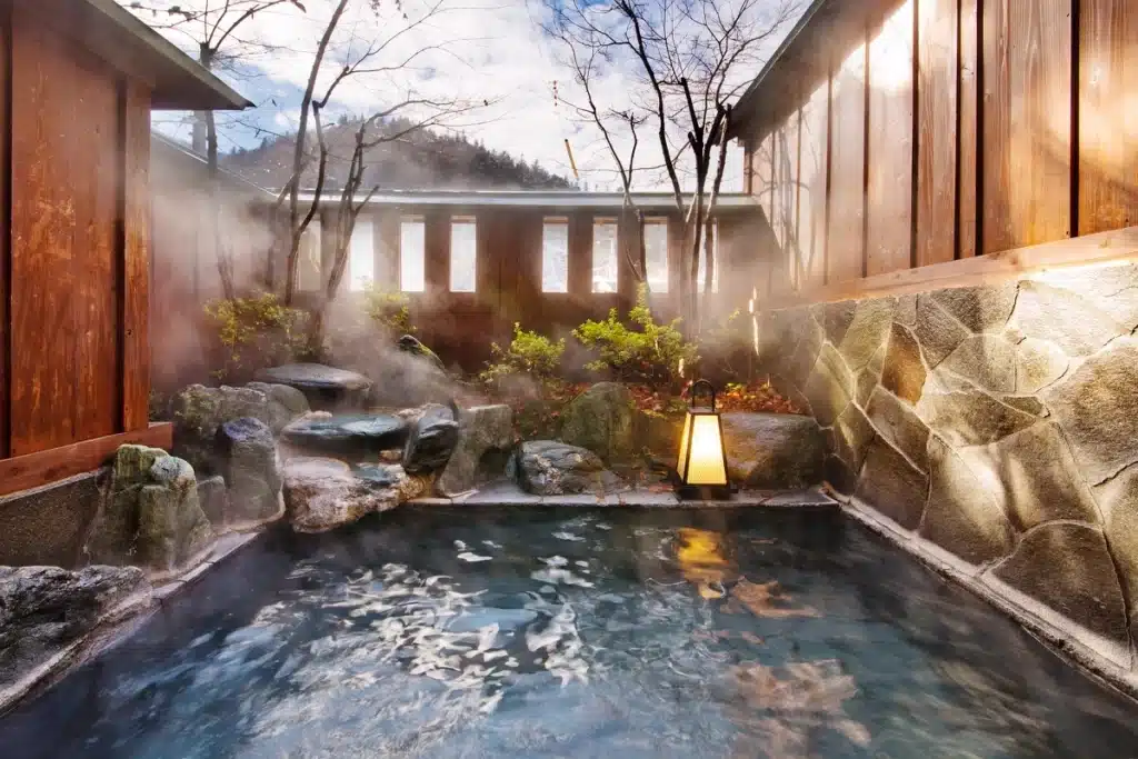 Steaming outdoor Japanese onsen bath surrounded by rocks, wooden walls, and trees, with a lantern by the waterβs edge on a cloudy day.