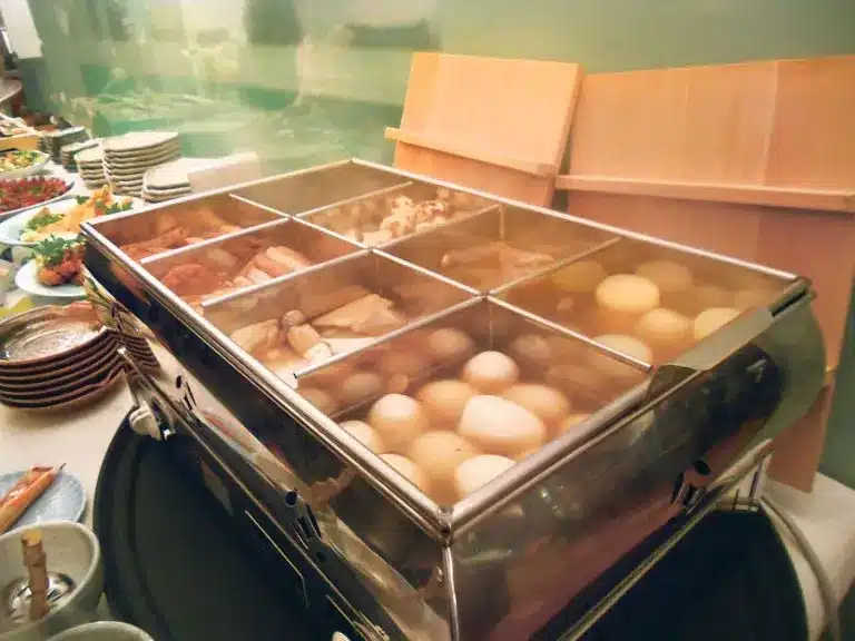 A stainless steel food warmer contains various simmered foods, including eggs and tofu—perfect for those wondering how to keep warm in winter—with plates and side dishes arranged on the table beside it.