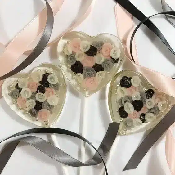 Three heart-shaped resin ornaments with pink, white, and dark flowers inside, placed on a white surface with pink and grey satin ribbons—perfect for a romantic white Valentine’s Day.