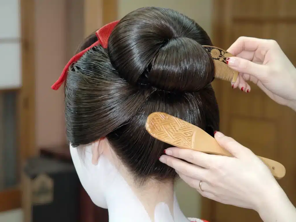 A person styles a Japanese geishaβs traditional Japanese updo with a wooden comb and hair tool; her neck is painted white.