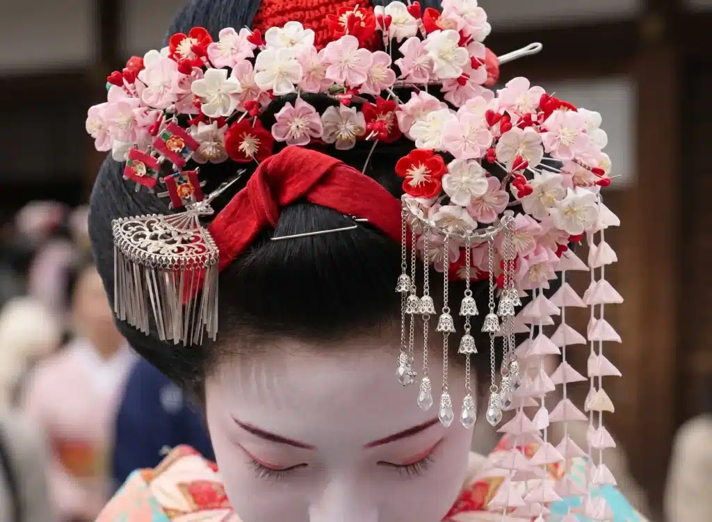 A Japanese geisha wearing traditional hair ornaments with intricate pink and red floral decorations, silver accessories, and white face make-up, looking downwards.