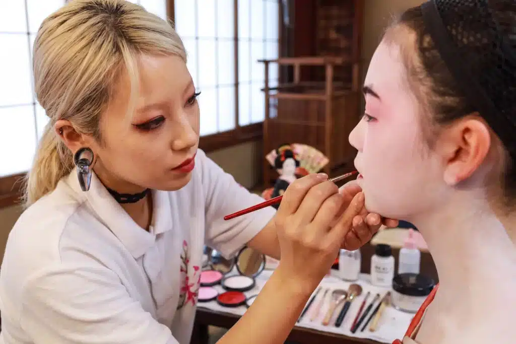 A make-up artist applies white and red make-up to a person's face in the style of a Japanese geisha, with cosmetics and brushes arranged on the table beside them.
