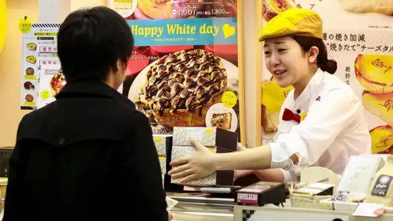 A shop worker in a yellow hat hands a boxed item to a customer across the counter, with dessert posters and white Valentine’s Day sweets visible in the background.
