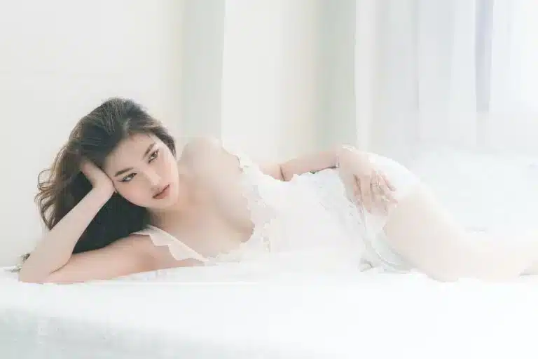 A woman in a white lace outfit is lying on her side on a white bed, resting her head on one hand and looking ahead. The bright, softly lit setting evokes the gentle charm of a white Valentine’s Day.