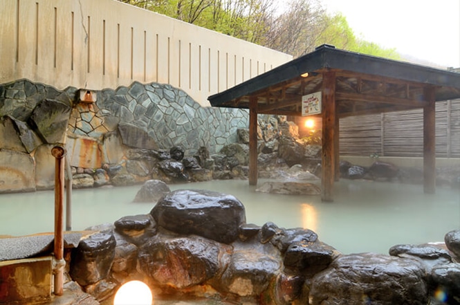 An outdoor hot spring bath with natural rock formations, steaming water, and a wooden shelter roof awaits you. The backdrop includes a stone wall and trees, providing the perfect serene escape.