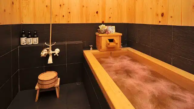 A traditional Japanese wooden soaking tub in a small bathroom with black tile walls, wooden accents, toiletries, and a stool with a wooden bucket—perfect for relaxing after enjoying a contactless spa promotion.