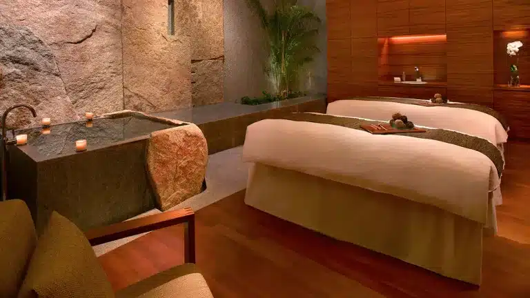 A tranquil spa room with two massage tables, stone wall accents, dim lighting, and a green plant in the corner. Candles are placed around for a calming ambiance, making it one of the most serene spots for massages in Singapore.