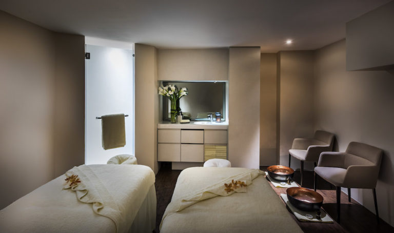 A serene spa room in Singapore features two massage tables, a sink with a mirrored cabinet, two chairs, and vases of calla lilies on the counter. Perfect for indulging in relaxing massages in Singapore.