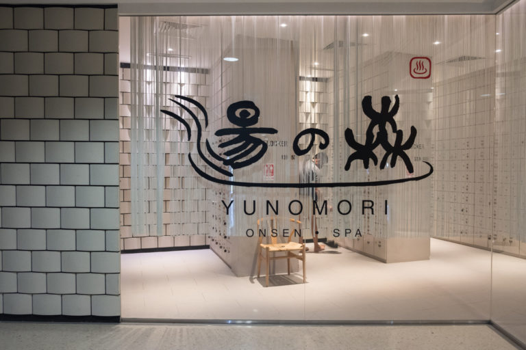 Yunomori Onsen & Spa entrance with a glass door, showcasing an interior with lockers and a wooden stool. A person can be seen inside, enjoying one of the best massages in Singapore.