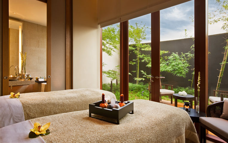 A serene spa room with two massage tables, a tray with oil bottles, and a view of a green outdoor garden through floor-to-ceiling glass doors offers one of the finest massages in Singapore.