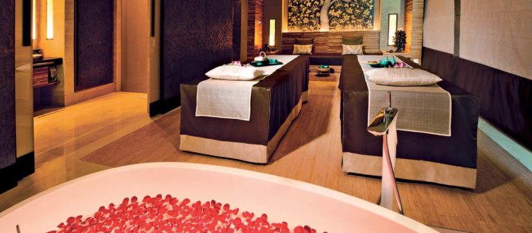 Luxurious spa room with two massage tables, neatly arranged towels, and rose petals in a bathtub in the foreground, perfect for indulging in massages in Singapore.