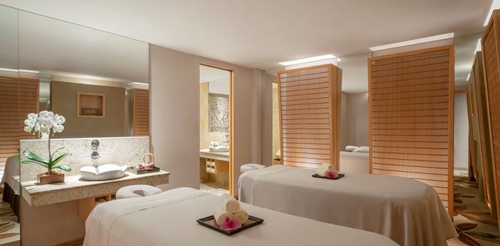 A serene spa room with two massage tables prepared with towels and flowers, dim lighting, and mirrored walls offers some of the most relaxing massages in Singapore.