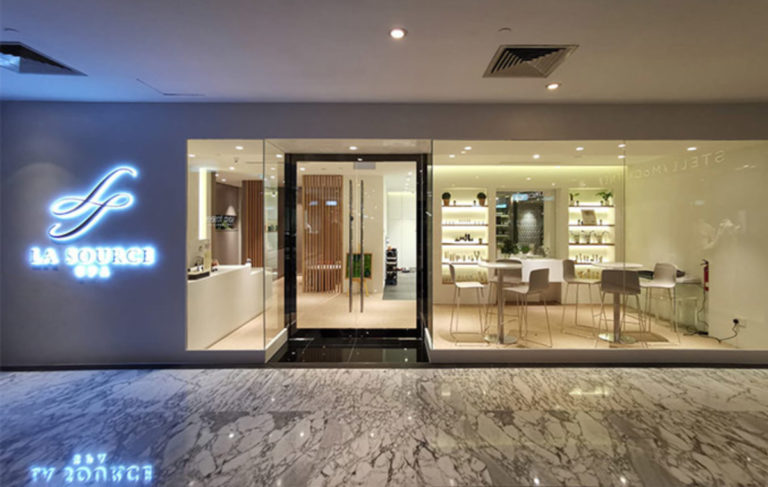 Modern spa entrance with glass doors, sleek interior design, illuminated signage "La Source Spa" on the left wall, and a marble floor in the foreground. Offering massages in Singapore, this spa features minimalist decor with light-colored furnishings.