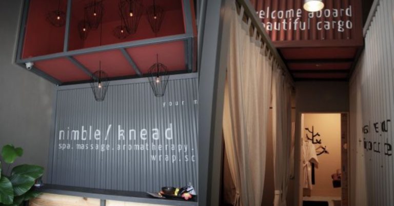 Entrance of Nimble/Knead spa with corrugated metal walls, hanging light fixtures, and a sign saying "Welcome aboard beautiful cargo." Corridor leads to treatment rooms with curtain partitions, offering some of the best massages in Singapore.