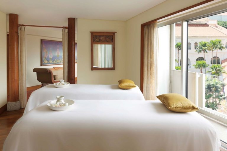 A serene spa room with two massage beds covered in white sheets, each adorned with a yellow pillow and a wooden tray bearing teacups, offering a perfect setting for massages in Singapore. The large window provides a calming view of the buildings and greenery outside.