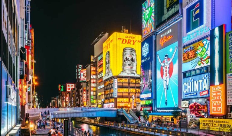 Night view of a busy street in a city with brightly lit billboards, including advertisements for Asahi beer and Glico, reflecting on a canal below. Pedestrians walk along the street and bridge.