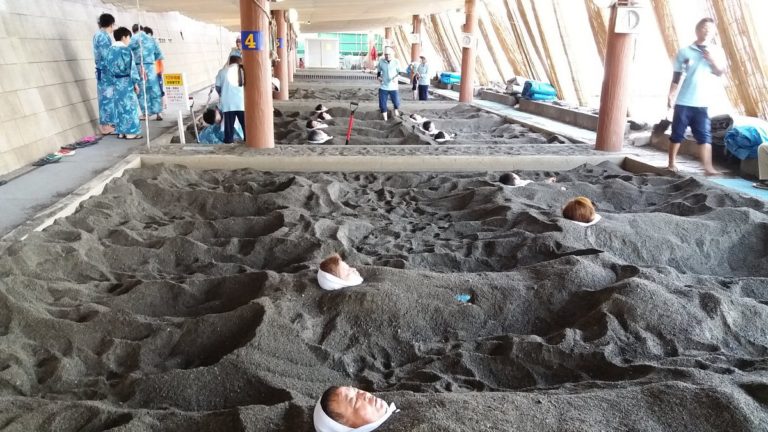 People are partially buried in sandy pits inside a facility, with only their heads sticking out, while others in blue robes walk and stand nearby, much like travelers making the most of their Japan Rail Pass in bustling stations.