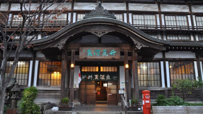 Front exterior of a traditional Japanese building with wooden architecture, a curved roof entrance, and a red post box on the side. Text above the door is in Japanese.