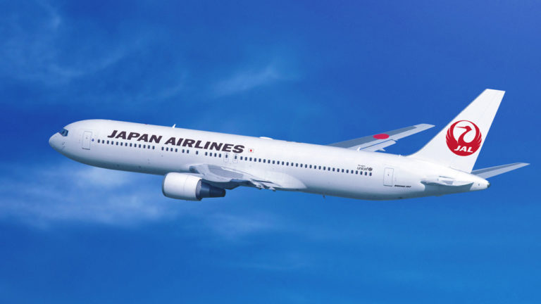 A Japan Airlines plane with a white body and red logo is flying in a clear blue sky