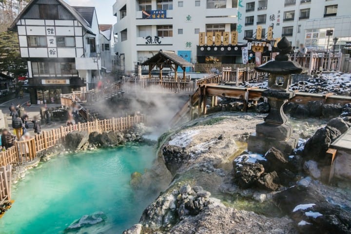A view of a hot spring in an urban area with steam rising from the water, surrounded by buildings and a wooden fence. Snow patches are visible on the rocks and ground. People, possibly travelers using a JR Pass, can be seen walking in the background.